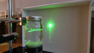A clear jar with a green laser shining through it and hitting a white board