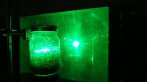 A jar with green liquid and green glitter being struck by a laser that transmits through to strike a white board